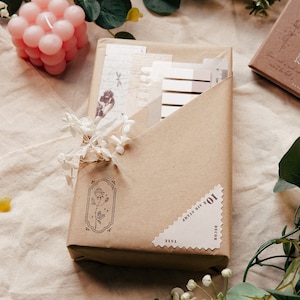 May include: A brown paper package with a white sticker that says "10% off any 3 items" and "Free Decor". The package is tied with a white ribbon and decorated with white flowers.