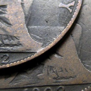 Op de afbeelding: Twee koperkleurige munten die op elkaar gestapeld zijn. De bovenste munt heeft het jaar 1902 gegraveerd op de rand. De onderste munt heeft een ontwerp van een persoonsprofiel en het jaar 1902 gegraveerd op de rand.