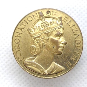 Puede incluir: Una moneda conmemorativa dorada que presenta el perfil de una persona coronada. La moneda está inscrita con "CORONATION OF ELIZABETH II". Tiene un pequeño agujero en la parte superior, lo que sugiere que podría usarse como colgante.