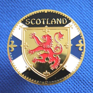 Puede incluir: Un emblema redondo y dorado con la palabra "SCOTLAND" en la parte superior. Presenta un león rojo rampante sobre un escudo dorado, rodeado por un borde negro y blanco con detalles azules. El fondo es de tela azul texturizada.