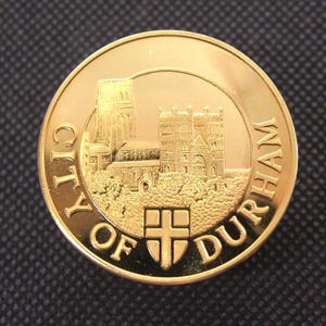 Puede incluir: Una moneda conmemorativa dorada de la ciudad de Durham. La moneda presenta una imagen en relieve de una catedral y las palabras "CITY OF DURHAM" alrededor del borde. Una cruz está en la parte inferior de la imagen de la catedral.