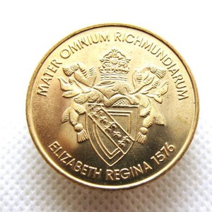 Puede incluir: Una moneda dorada con texto en relieve y un escudo de armas. La moneda presenta el texto "MATER OMNIUM RICHMUNDIARUM" y "ELIZABETH REGINA 1576". La moneda tiene un diseño detallado con una corona y un escudo.