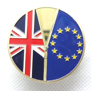 Puede incluir: Un pin redondo con borde dorado que muestra la bandera del Reino Unido a la izquierda y la bandera de la Unión Europea a la derecha. Una cremallera dorada separa ambas banderas. La bandera de la UE tiene estrellas amarillas sobre fondo azul.