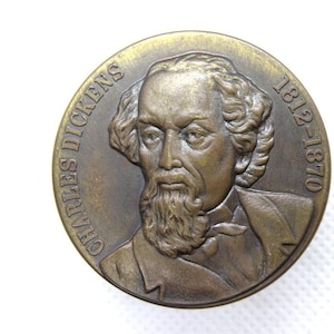 Puede incluir: Medalla de bronce con un retrato de Charles Dickens. La inscripción "CHARLES DICKENS" está en la parte superior, con las fechas "1812-1870" a la derecha. El retrato muestra a un hombre con barba y chaqueta.