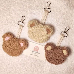 May include: Three brown and cream fuzzy bear keychains with a white ribbon attached to each keychain that says "with love". The keychains are on a white fluffy surface. A small white card with a heart and the text "HARIKA THINGS GIFT WORKSHOP" is in the centre of the image.