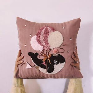 Könnte beinhalten: Ein rosa Kissen mit einem Wal, der drei rosa und weiße Ballons festhält. Der Wal trägt eine Krone und eine Schleife. Das Kissen ist mit Strasssteinen verziert.