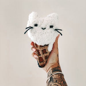 Könnte beinhalten: Ein weißer, flauschiger Eiswaffel in Katzenform mit schwarzen Augen und Schnurrhaaren. Die Waffel ist braun und hat ein Waffelmuster.
