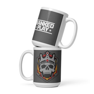 May include: Two white ceramic mugs with different designs. The top mug has a gray background with the text "RANKED PLAY" in white. The bottom mug has a gray background with a stylized skull wearing a crown and surrounded by yellow and red flames.