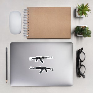 May include: Two black AK-47 rifle stickers on a laptop. The stickers are detailed with realistic features. The laptop is on a light-colored surface, with a notepad, mouse, pencil, glasses, and small potted plants nearby.