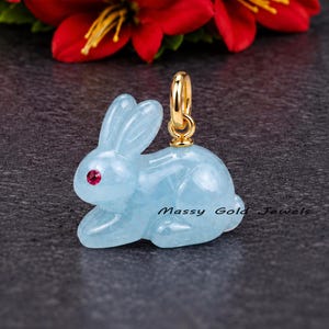 May include: A light blue carved gemstone rabbit pendant with a small red stone eye and a gold bail. The pendant features the text Massy Gold Jewels and is positioned in front of a cluster of vibrant red and white lily flowers.