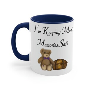 May include: White ceramic mug with a blue rim and handle. The mug features a cartoon teddy bear holding a treasure chest with the words "Memories Safe" and "I'm Keeping Mom's" printed on the mug.