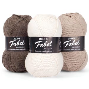 Puede incluir: Tres ovillos de lana Drops Fabel Uni Colour, lana para calcetines tratada con superlavado. El hilo está disponible en tres colores: marrón, blanco y beige. Cada ovillo tiene una etiqueta negra con el nombre de la marca y los detalles del producto.