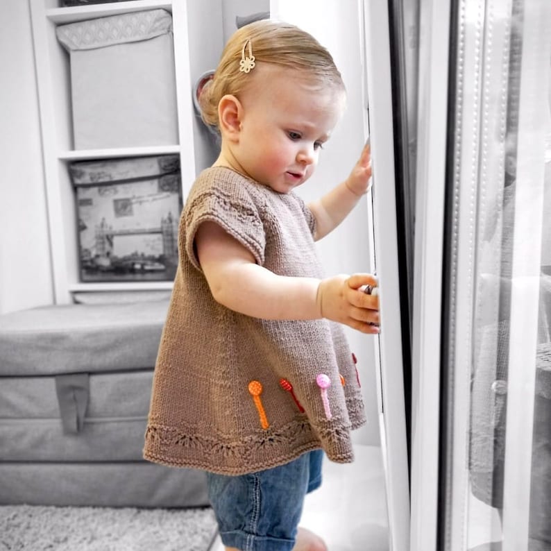 K&ouml;nnte beinhalten: Ein kleines Kind tr&auml;gt einen braunen Strickpullover mit bunten Akzenten und blaue Jeans. Der Pullover hat einen gewellten Saum und kurze &Auml;rmel.