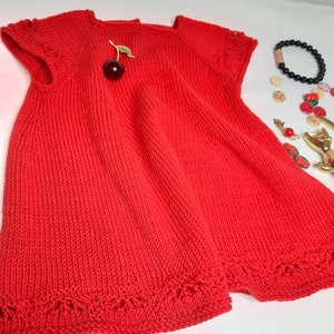 K&ouml;nnte beinhalten: Ein rotes Strickkleid mit kurzen &Auml;rmeln und einem zarten Spitzenbesatz am Saum. Das Kleid hat vorne eine Brosche in Form einer Kirsche.