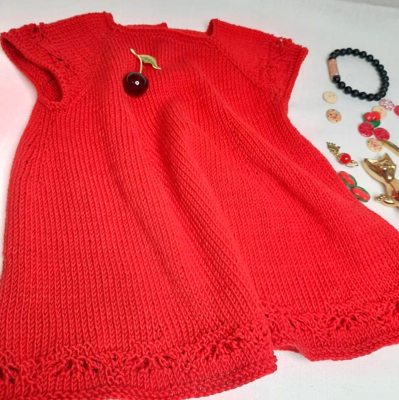 K&ouml;nnte beinhalten: Ein rotes Strickkleid mit kurzen &Auml;rmeln und einem zarten Spitzenbesatz am Saum. Das Kleid hat vorne eine Brosche in Form einer Kirsche.