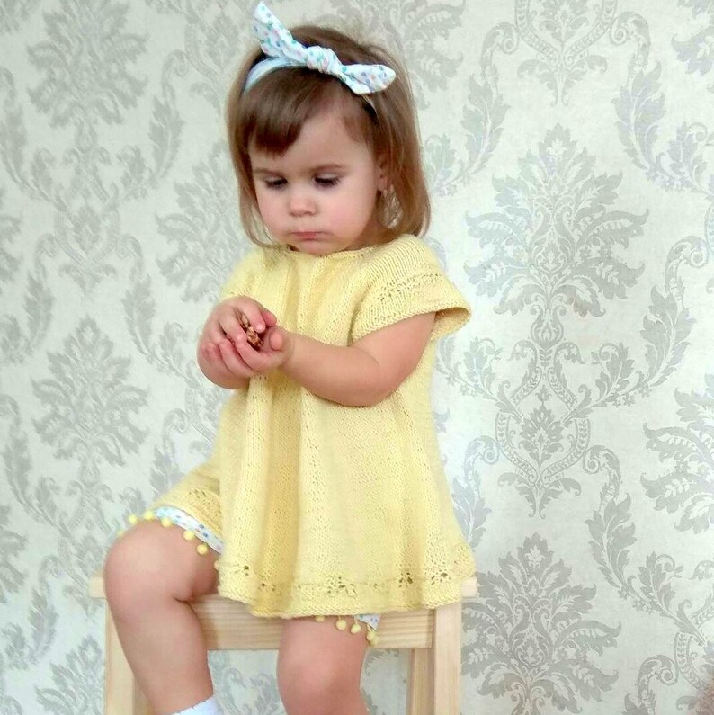 K&ouml;nnte beinhalten: Ein kleines M&auml;dchen tr&auml;gt ein gelbes Strickkleid mit einem wei&szlig;en Stirnband. Das Kleid hat einen gewellten Saum und Bommeln an den &Auml;rmeln.