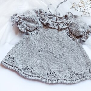 May include: A gray knitted baby dress with a ruffled collar and short sleeves. The dress has a delicate lace pattern around the neckline and hem.