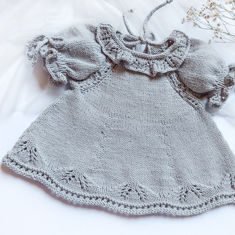 May include: A gray knitted baby dress with a ruffled collar and short sleeves. The dress has a delicate lace pattern around the neckline and hem.