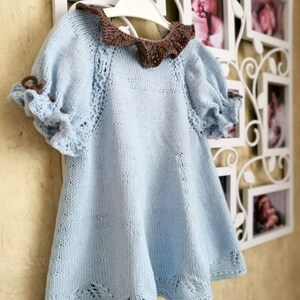May include: A light blue knitted baby dress with a brown crocheted collar and ruffled sleeves.