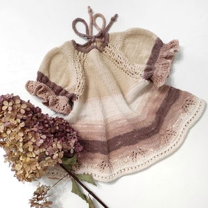May include: A knitted baby dress with a gradient of beige, pink, and brown. The dress has a ruffled neckline and sleeves.