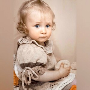 May include: A young child wearing a knitted beige dress with a white lace trim and brown stripes on the sleeves. The dress has a ruffled collar and a tie at the wrist.