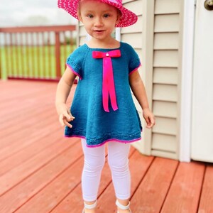 K&ouml;nnte beinhalten: Ein junges M&auml;dchen tr&auml;gt ein t&uuml;rkisfarbenes Strickkleid mit einer rosa Schleife und einem rosa Hut. Sie steht auf einem Holzdeck.