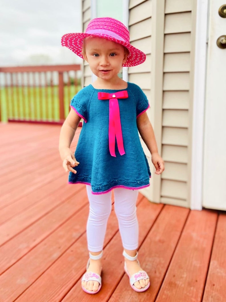 K&ouml;nnte beinhalten: Ein junges M&auml;dchen tr&auml;gt ein t&uuml;rkisfarbenes Strickkleid mit einer rosa Schleife und einem rosa Hut. Sie steht auf einem Holzdeck.