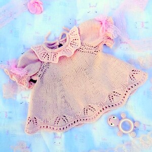 May include: A delicate pink knitted baby dress with a lace collar and short sleeves. The dress is laid out on a light blue fabric with a rabbit print.