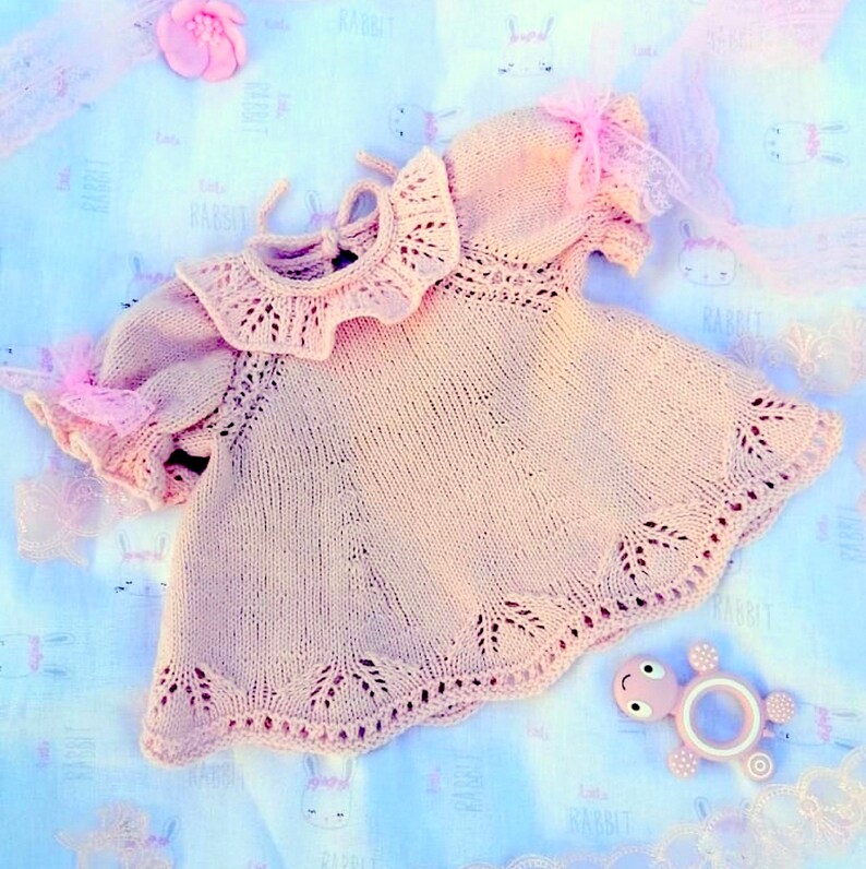 May include: A delicate pink knitted baby dress with a lace collar and short sleeves. The dress is laid out on a light blue fabric with a rabbit print.