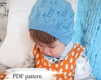 May include: A baby wearing a light blue knitted hat with a cable knit pattern. The hat has a brim and covers the baby's ears. The baby is wearing a brown and white polka dot shirt. The image includes the text "PDF pattern".
