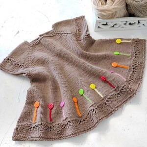 K&ouml;nnte beinhalten: Ein braunes gestricktes Babykleid mit bunten Kn&ouml;pfen und Schleifenakzenten.