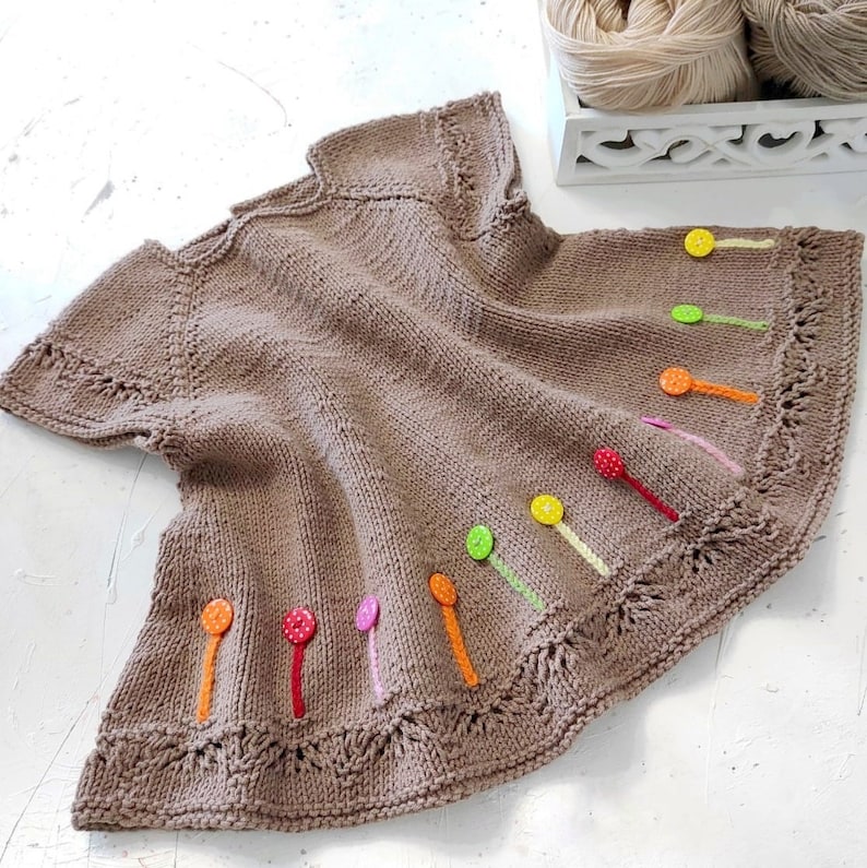 K&ouml;nnte beinhalten: Ein braunes gestricktes Babykleid mit bunten Kn&ouml;pfen und Schleifenakzenten.