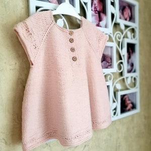 K&ouml;nnte beinhalten: Ein rosa gestricktes Babykleid mit kurzen &Auml;rmeln und Knopfleiste. Das Kleid hat einen zarten Spitzenbesatz am Ausschnitt und Saum.