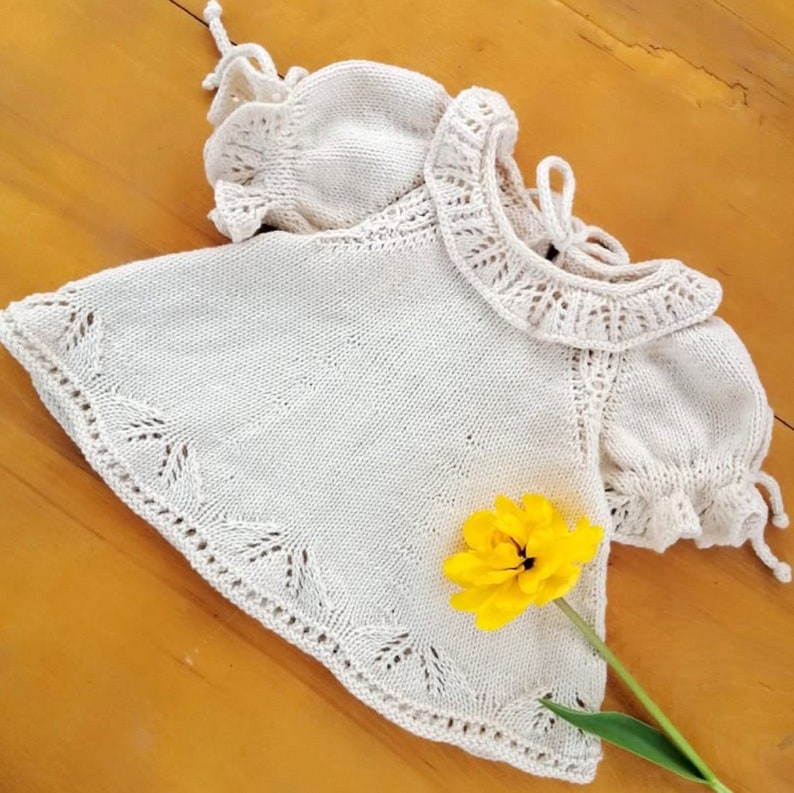 May include: A white knitted baby dress with a lace trim and ruffled sleeves. The dress is laid on a wooden surface with a yellow flower.