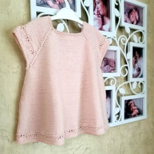 K&ouml;nnte beinhalten: Ein hellrosa gestricktes Babykleid mit kurzen &Auml;rmeln und einer zarten Spitzenborte am Ausschnitt und Saum.