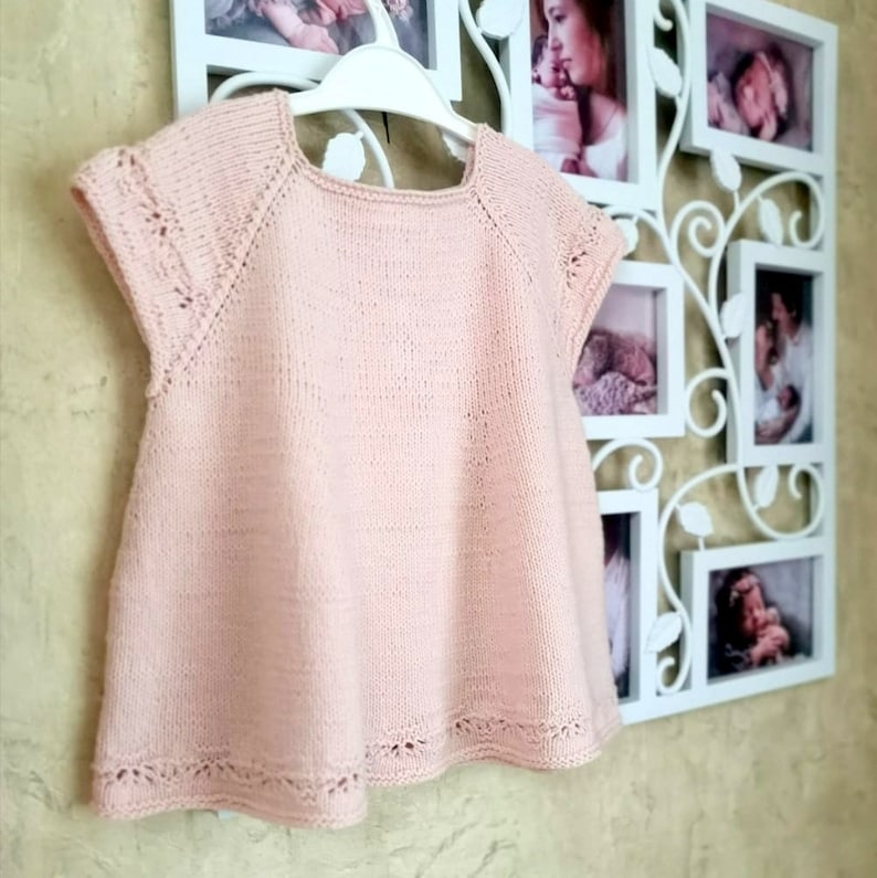 K&ouml;nnte beinhalten: Ein hellrosa gestricktes Babykleid mit kurzen &Auml;rmeln und einer zarten Spitzenborte am Ausschnitt und Saum.