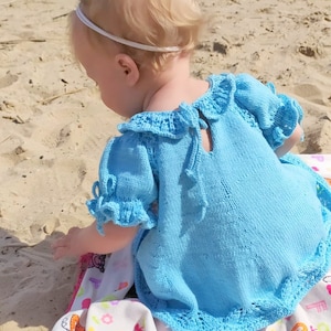 May include: A light blue knitted baby dress with a ruffled collar and sleeves. The dress has a tie at the back and a crocheted trim.