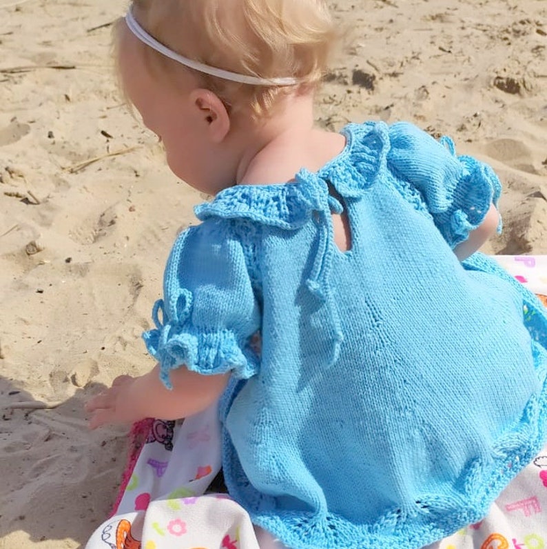 May include: A light blue knitted baby dress with a ruffled collar and sleeves. The dress has a tie at the back and a crocheted trim.