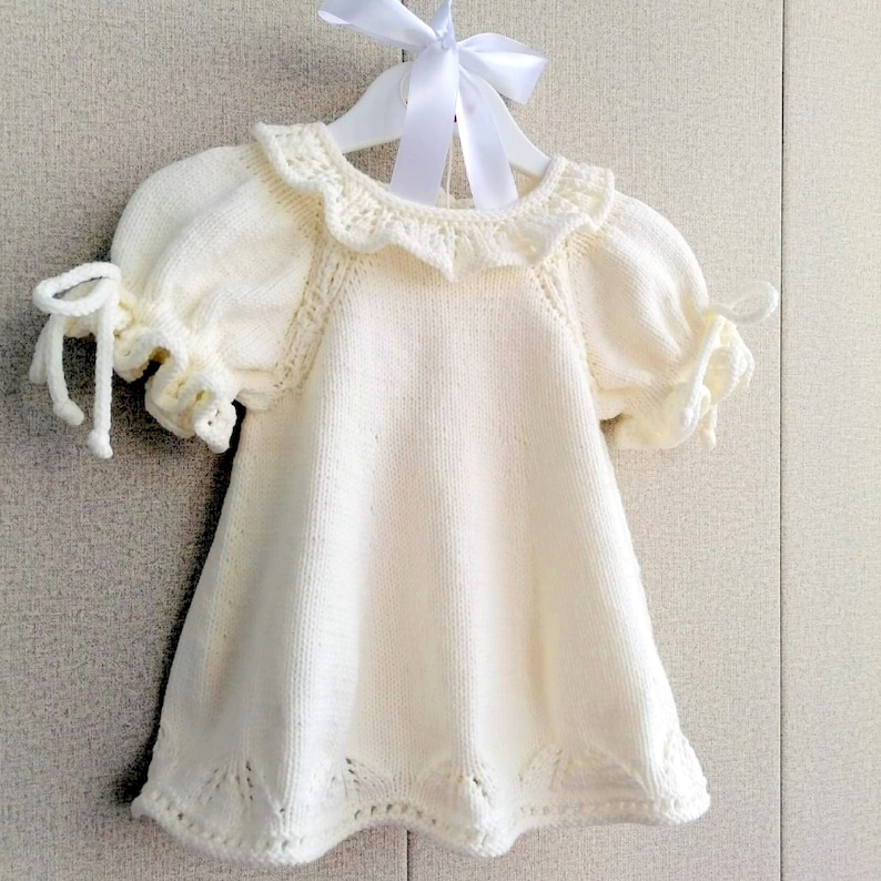 May include: A white knitted baby dress with a ruffled collar and short sleeves. The dress has a delicate lace pattern and is perfect for a special occasion.