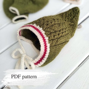 Puede incluir: Un gorro de bebé de punto verde con ribete blanco y rojo. El gorro tiene un lazo en la parte posterior. Patrón PDF.
