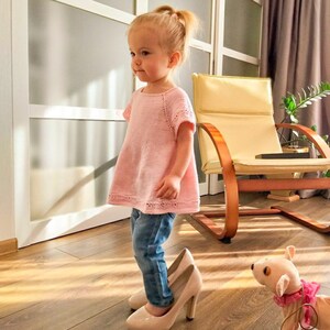 K&ouml;nnte beinhalten: Ein junges M&auml;dchen in einem rosa Strickoberteil und blauen Jeans steht auf einem Paar hochhackiger Schuhe. Sie schaut nach links aus dem Bild. Ein kleines Stofftier liegt rechts neben dem M&auml;dchen auf dem Boden.