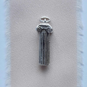 Puede incluir: Un charm de color plateado con forma de columna clásica. El charm presenta acanaladuras detalladas en la columna y un capitel decorativo con diseños en forma de voluta. En la parte superior hay un pequeño lazo para sujetarlo a una cadena o pulsera.