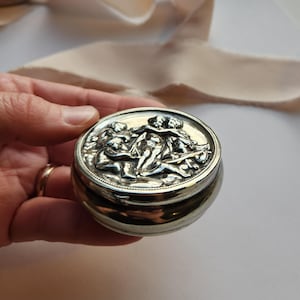 May include: Silver-toned, round trinket box with a detailed relief of classical figures on the lid. The box is held in a hand, showcasing its size and design. The background includes a soft, neutral-toned fabric.
