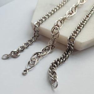 Könnte beinhalten: Drei silberfarbene Ketten mit unterschiedlichen Glieddesigns werden vor einem weißen Hintergrund gezeigt. Die Ketten umfassen eine Panzerkette, eine Seilkette und eine Kette mit ovalen Gliedern. Jede Kette hat eine Schlaufe am Ende.