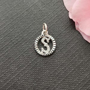May include: A silver pendant featuring the letter "S" within a circular frame. The letter and frame are adorned with small, sparkling details. The pendant is designed for use as a necklace charm or bracelet bead.