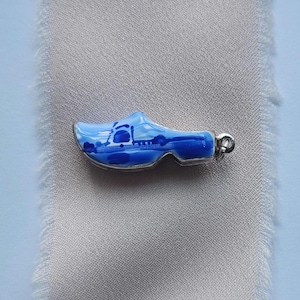 May include: A miniature blue and white wooden clog charm with a silver-coloured metal loop. The clog features a painted landscape scene. The charm is a traditional Dutch souvenir.