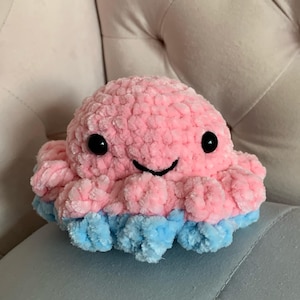 Peut inclure: Une peluche pieuvre rose avec un visage souriant et des yeux noirs. Les tentacules sont bleu clair. Le jouet est fait d'une matière douce et texturée.