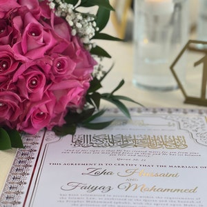 May include: A white wedding ceremony card with gold accents and Arabic calligraphy. The card states "This agreement is to certify that the marriage of Zoha Hussaini and Faiyaz Mohammed." The date of the ceremony is January 28, 2023.
