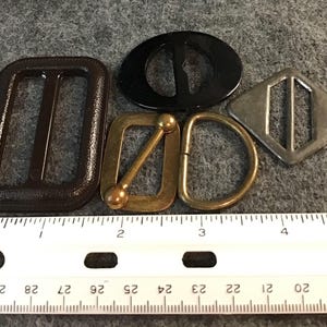 May include: A collection of five assorted buckles including a large dark brown rectangular buckle, a black oval buckle, a brass rectangular buckle with a center bar, a brass D-ring, and a silver-toned diamond-shaped buckle, shown with a ruler.