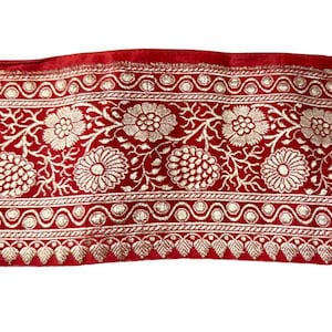 May include: A section of red fabric with an ornate gold floral pattern. The design features large, detailed flowers, vines, and decorative borders. The fabric has a velvet-like texture and is likely used for decorative purposes.